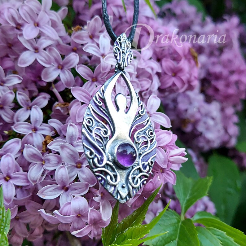 May include: A silver pendant featuring a stylized female figure with an amethyst gemstone in the center. The pendant is surrounded by intricate floral designs and hangs from a black cord.