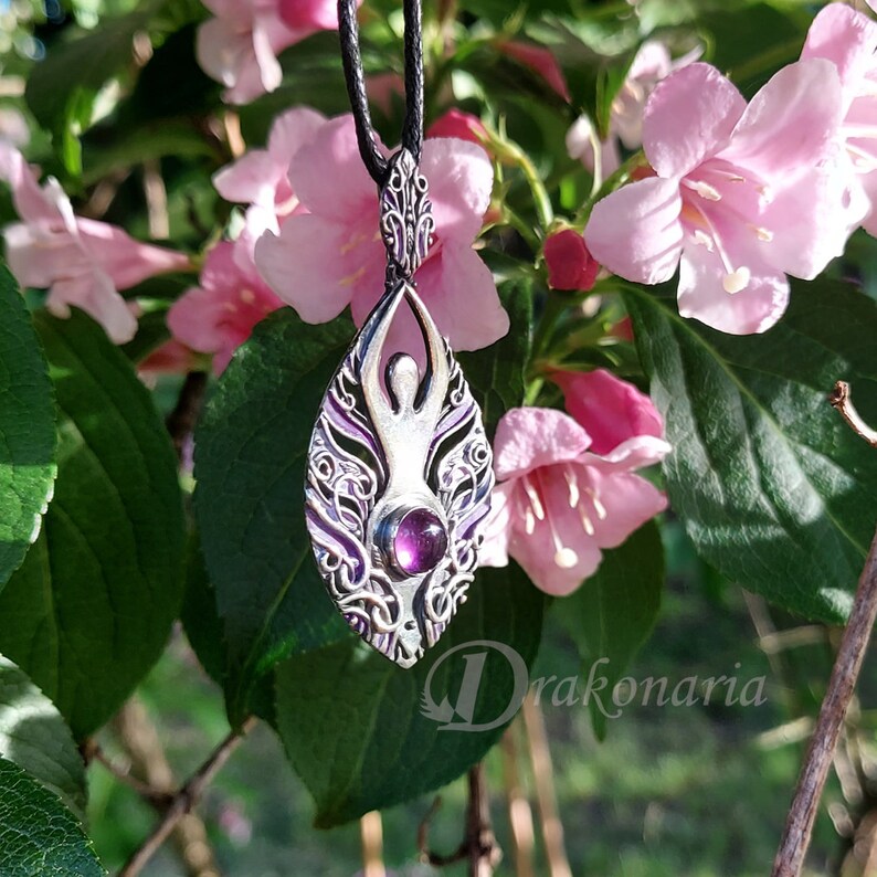 May include: A silver pendant with an intricate design featuring a stylized human figure and a purple gemstone. The pendant is hanging from a black cord and is surrounded by pink flowers.