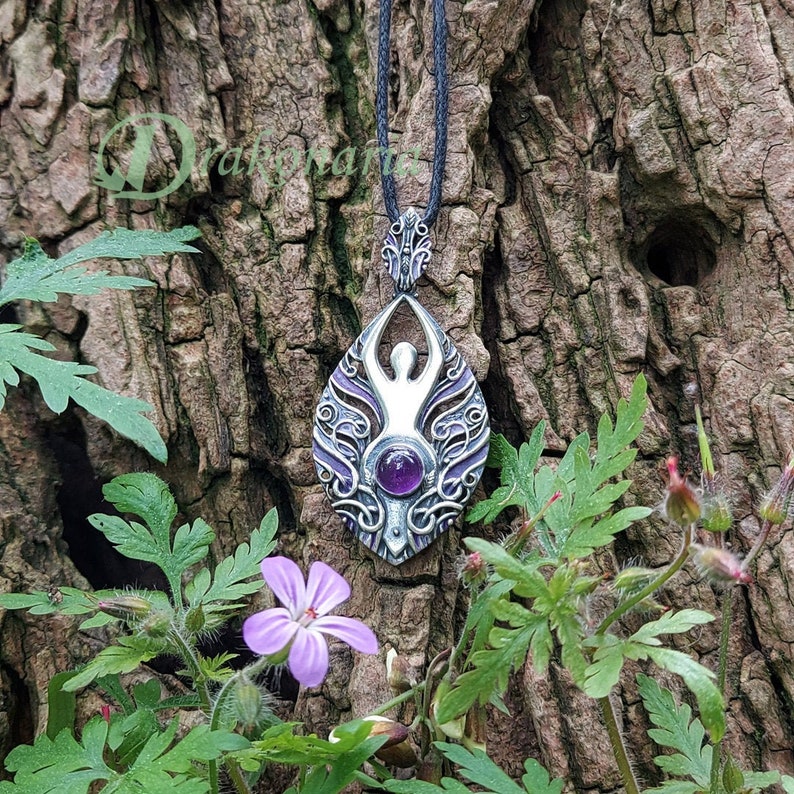 May include: Silver pendant with an intricate design featuring a stylized human figure and a purple amethyst gemstone. The pendant is hanging from a black cord and is surrounded by green foliage.