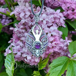 May include: A silver pendant with an intricate design featuring a stylized human figure and a purple amethyst gemstone. The pendant is hanging from a black cord and is surrounded by purple flowers.