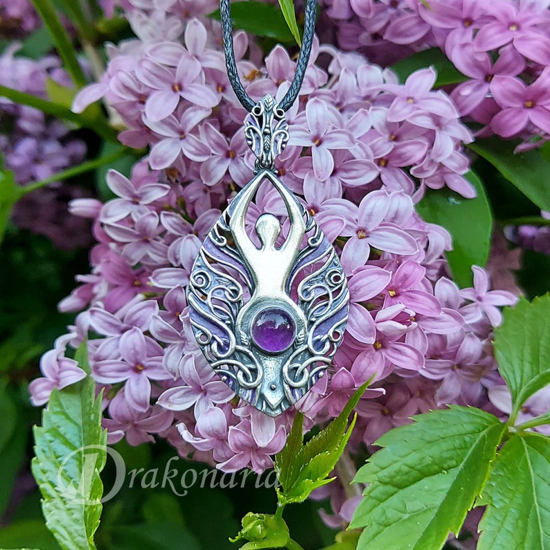 May include: A silver pendant with an intricate design featuring a stylized human figure and a purple amethyst gemstone. The pendant is hanging from a black cord and is surrounded by purple flowers.