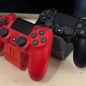 Peut inclure: Deux manettes de jeu PlayStation 4, l'une rouge et l'autre noire, sont présentées sur un support. Les manettes sont toutes les deux dans la même position, avec les boutons face avant.