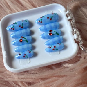 May include: A set of ten blue and white gingham press-on nails with red cherry designs. The nails are arranged in a white plastic case.