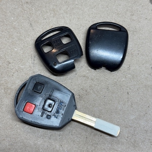 Puede incluir: Llave de coche negra con una hoja plateada, un botón de pánico rojo y botones con las etiquetas "LOCK" y "UNLOCK". La llave va acompañada de dos carcasas negras para el mando.