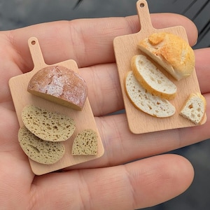 Op de afbeelding: Twee miniatuur houten snijplanken met sneetjes brood. Een plank heeft een heel brood en meerdere sneetjes brood. De andere plank heeft meerdere sneetjes brood.