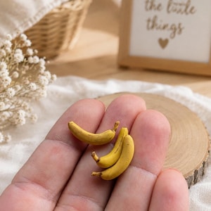 Op de afbeelding: Drie miniatuur bananen in het geel met bruine steeltjes, ontworpen als realistisch fruit. Deze kleine replica's hebben een gebogen vorm en een gladde, matte textuur, met gedetailleerde schaduwen die het uiterlijk van rijpe bananen nabootsen.