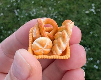 May include: A small orange basket filled with miniature baked goods, including a pretzel, a loaf of bread, and two rolls. The basket is held in a person's hand.