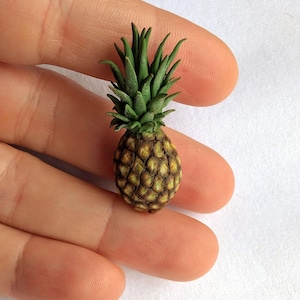 May include: A miniature pineapple with a textured, brown exterior and a crown of green leaves. The small fruit is held in a hand, showcasing its size. The pineapple is a detailed, handcrafted item.