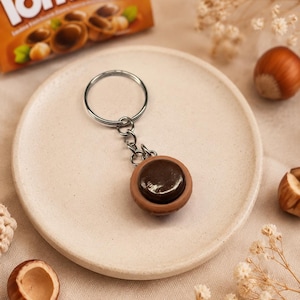 Può includere: Un portachiavi con una replica in miniatura di un dolcetto Toffifee, composto da una coppetta di caramello marrone chiaro riempita con un centro di cioccolato marrone scuro, fissato a un anello di metallo argentato da una catenella.