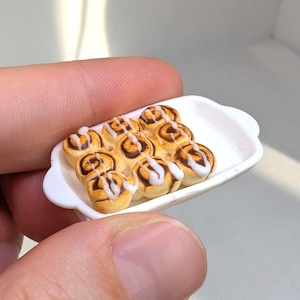 Könnte beinhalten: Miniatur-Zimtschnecken mit weißem Zuckerguss auf einem kleinen weißen Tablett. Die Brötchen sind goldbraun mit einem Wirbelmuster und einem Guss aus weißem Zuckerguss. Das Tablett wird von den Fingern einer Person gehalten.
