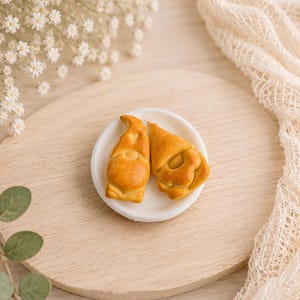 Könnte beinhalten: Zwei goldbraune Zwergenfiguren im Miniaturformat mit spitzen Hüten und runden Körpern, die nebeneinander auf einem kleinen, runden weißen Teller liegen. Die Zwerge haben glatte, gebacken wirkende Oberflächen mit feinen Details.