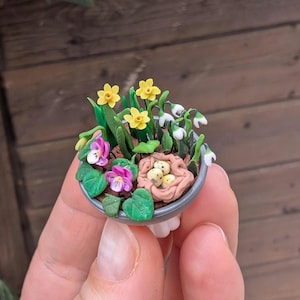Könnte beinhalten: Ein kleiner Blumentopf aus Ton mit einem Nest aus gelben Eiern, umgeben von gelben Narzissen, weißen Schneeglöckchen und violetten Veilchen.