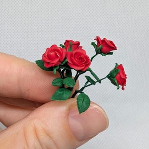Puede incluir: Un pequeño ramo de rosas rojas con hojas verdes. Las rosas están hechas de arcilla polimérica y tienen aproximadamente el tamaño de una moneda de 10 céntimos.