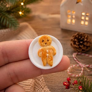 Könnte beinhalten: Eine Miniatur-Lebkuchenfigur mit goldbrauner Backtextur, die zwei dunkle Augen und zwei kleine weiße Knöpfe am Oberkörper aufweist und auf einem kleinen, schlichten weißen runden Teller mit strukturiertem Rand zentriert ist.