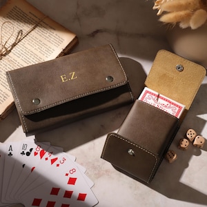 May include: Two brown leather card cases, one open with a deck of cards and the other closed with gold initials. A vintage book and dice are also present. The card cases have a snap closure.