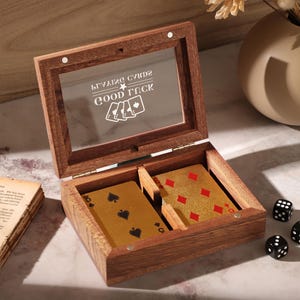 Könnte beinhalten: Holzspielkartenbox mit transparentem Deckel, auf dem der Text "GOOD LUCK" und Kartensymbole zu sehen sind. Die Box enthält zwei goldfarbene Spielkarten und wird von drei schwarzen Würfeln begleitet. Ein Stapel alter Bücher und eine beige Vase befinden sich im Hintergrund.
