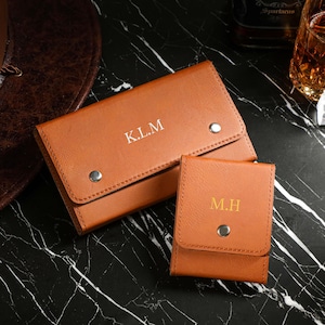 May include: Two brown leather cases, one rectangular and one square, with silver snap closures. The larger case has the initials "K.L.M." in white, and the smaller case has "M.H." in gold. The items are on a black and white marble surface.