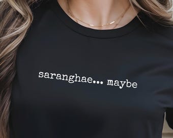 Puede incluir: Camiseta negra con el texto blanco "saranghae... maybe". La camiseta tiene mangas cortas y cuello redondo. La modelo lleva vaqueros azules.