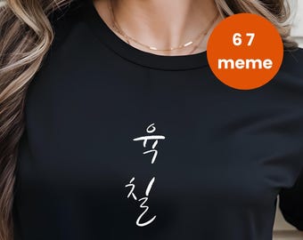 Puede incluir: Camiseta negra con escritura coreana blanca y una pegatina redonda naranja que dice "67 meme". La camiseta tiene mangas cortas y cuello redondo. La modelo lleva vaqueros azules.