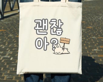 Puede incluir: Un bolso tote de color crema con texto coreano negro y un conejo de dibujos animados sosteniendo un letrero. El bolso lo lleva una persona con jeans azules y botas marrones.