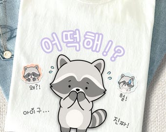 Puede incluir: Una camiseta blanca con una ilustración central de un mapache de dibujos animados con las patas sobre la boca. La camiseta incluye texto en coreano en morado claro y negro, con gráficos de mapaches más pequeños a los lados.