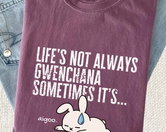 Puede incluir: Una camiseta color baya con el texto blanco "LIFE'S NOT ALWAYS GWENCHANA SOMETIMES IT'S..." sobre un gráfico de un conejo blanco con una gota de sudor azul y la palabra "aigoo." impresa en blanco en la parte inferior izquierda del gráfico.