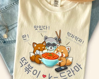 Puede incluir: Una camiseta de color marfil con un gráfico de tres animales de dibujos animados comiendo un tazón de tteokbokki rojo. El diseño incluye texto en coreano, las frases masitda, jjan, meokja meokja y el texto tteokbokki + k-dramas con un pequeño corazón rojo.