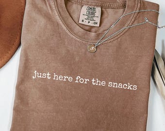 Puede incluir: Camiseta color espresso con la frase "just here for the snacks" en blanco. Un collar plateado con un colgante descansa sobre la camiseta. Un sombrero marrón es visible en la esquina superior izquierda.