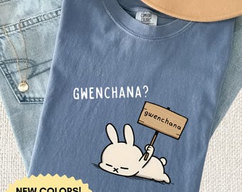 Puede incluir: Una camiseta de color azul vaquero con un gráfico de un conejo blanco tumbado que sostiene un cartel de madera con la palabra "gwenchana". Sobre el conejo, el texto "GWENCHANA?" está impreso en blanco. La camiseta tiene cuello redondo.