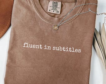 Puede incluir: Una camiseta color espresso con cuello redondo y el texto blanco "fluent in subtitles" impreso en el pecho. La prenda cuenta con una etiqueta de la marca Comfort Colors en el cuello interior, indicando una talla small.