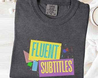 Puede incluir: Camiseta de color grafito con las palabras "FLUENT SUBTITLES" en un diseño retro colorido. La camiseta tiene cuello redondo y una etiqueta "Comfort Colors". La camiseta está doblada y colocada sobre una superficie blanca.