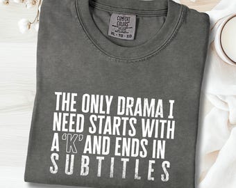 Puede incluir: Una camiseta de color pimienta con el texto blanco "THE ONLY DRAMA I NEED STARTS WITH A 'K' AND ENDS IN SUBTITLES". La camiseta está doblada y tiene una etiqueta Comfort Colors. Una pequeña taza blanca y flores en el fondo.
