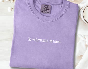 Puede incluir: Camiseta color orquídea con el texto "k-drama mama" en blanco. La camiseta es de la marca Comfort Colors, talla XL-TG-EG. La camiseta está doblada y colocada sobre una superficie clara, con una pequeña vela blanca y detalles florales.