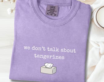 Puede incluir: Camiseta color orquídea con el texto "we don't talk about tangerines" y un gráfico de caja de pañuelos. La camiseta es de la marca Comfort Colors, talla XL-TG-EG. La camiseta está doblada sobre una superficie blanca.