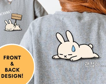 Puede incluir: Sudadera gris deportiva con diseño frontal y trasero. La parte delantera presenta un conejito de dibujos animados sosteniendo un letrero que dice "gwenchana?". La parte trasera tiene un conejito con una gota de sudor y texto coreano.