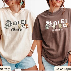May include: Ivory and espresso-colored t-shirts with a Korean text design and cute animal illustrations. The text reads "HWAITING!" with a raccoon, rabbit, bear, and red panda. The shirts are short-sleeved and have a relaxed fit.