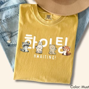 May include: Mustard-colored t-shirt with the Korean word "HWAITING!" and cute animal illustrations. The shirt is folded on top of a pair of blue jeans. A tan hat is visible in the background.