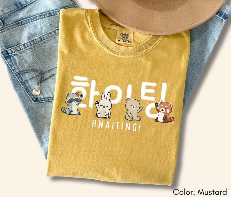 May include: Mustard-colored t-shirt with the Korean word "HWAITING!" and cute animal illustrations. The shirt is folded on top of a pair of blue jeans. A tan hat is visible in the background.