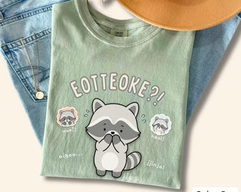 Puede incluir: Una camiseta color baya con un gráfico central de un mapache de dibujos animados con el texto EOTTEOKE?! encima. Ilustraciones más pequeñas de mapaches aparecen a los lados con el texto wae?!, heol!, aigoo... y jjinja! impreso en blanco.
