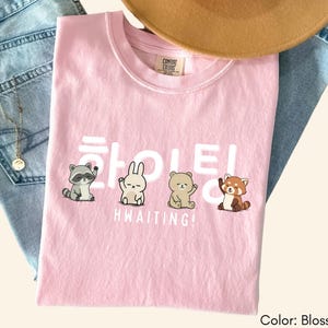 May include: A light pink t-shirt with the text "HWAITING!" in white, along with illustrations of a raccoon, rabbit, bear, and red panda. The shirt is folded on top of a pair of blue jeans, with a gold necklace and a brown hat.
