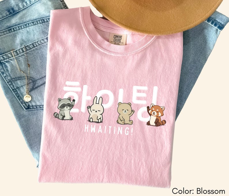 May include: A light pink t-shirt with the text "HWAITING!" in white, along with illustrations of a raccoon, rabbit, bear, and red panda. The shirt is folded on top of a pair of blue jeans, with a gold necklace and a brown hat.