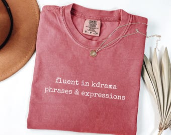 Puede incluir: Camiseta de color rojo-marrón con el texto "fluent in kdrama phrases & expressions" en blanco. Un collar dorado con un colgante descansa sobre la camiseta. Un sombrero marrón es visible en el fondo.