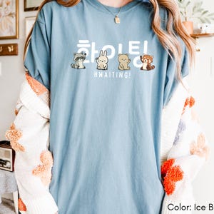 May include: Ice blue t-shirt with the word "Hwaiting!" in white Korean characters and English text. The shirt features cartoon illustrations of a raccoon, rabbit, bear, and red panda. The shirt is a light blue color.