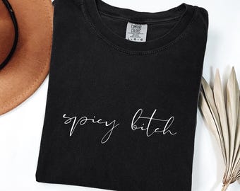 Puede incluir: Camiseta negra con las palabras "spicy bitch" en escritura cursiva blanca. La camiseta está doblada y tiene una etiqueta Comfort Colors. Un sombrero marrón y una planta seca están en el fondo.