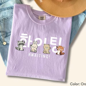 May include: Orchid-colored t-shirt with the word "HWAITING!" in white, along with cartoon animal illustrations. The shirt is displayed on a pair of blue jeans, with a gold necklace and a brown hat in the background.