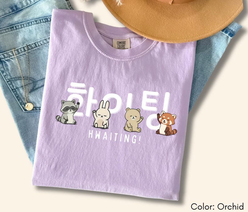 May include: Orchid-colored t-shirt with the word "HWAITING!" in white, along with cartoon animal illustrations. The shirt is displayed on a pair of blue jeans, with a gold necklace and a brown hat in the background.