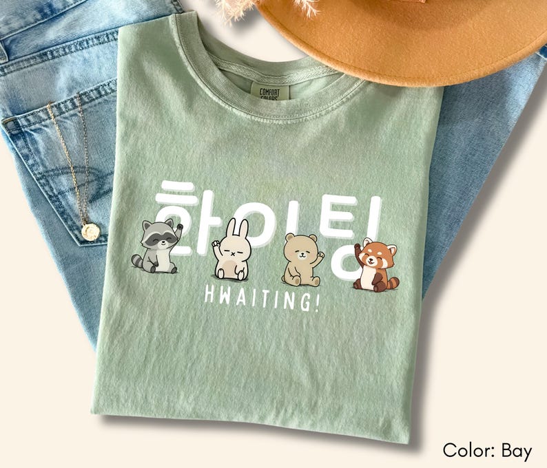 May include: A bay-coloured t-shirt with a graphic featuring Korean text and the English word "HWAITING!" below. The graphic includes illustrations of a raccoon, a rabbit, a bear, and a red panda. The shirt is laid on top of blue jeans.