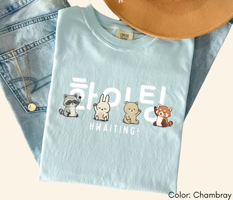 May include: A light blue t-shirt with the word "HWAITING!" in white, along with illustrations of a raccoon, a bunny, a bear, and a red panda. The shirt is made of a soft material and is a chambray color.