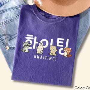 May include: A grape-colored t-shirt with a graphic featuring cartoon animals and Korean text. The text reads "HWAITING!" with a raccoon, rabbit, bear, and red panda. The shirt is folded on top of blue jeans and a brown hat.