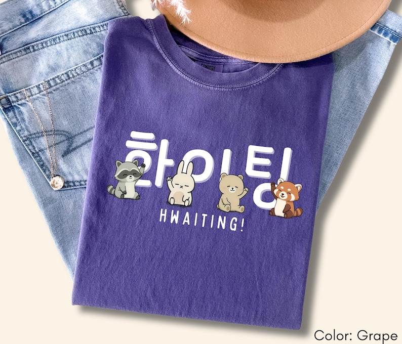 May include: A grape-colored t-shirt with a graphic featuring cartoon animals and Korean text. The text reads "HWAITING!" with a raccoon, rabbit, bear, and red panda. The shirt is folded on top of blue jeans and a brown hat.